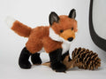 Plush red fox standing beside pinecones and autumn leaves